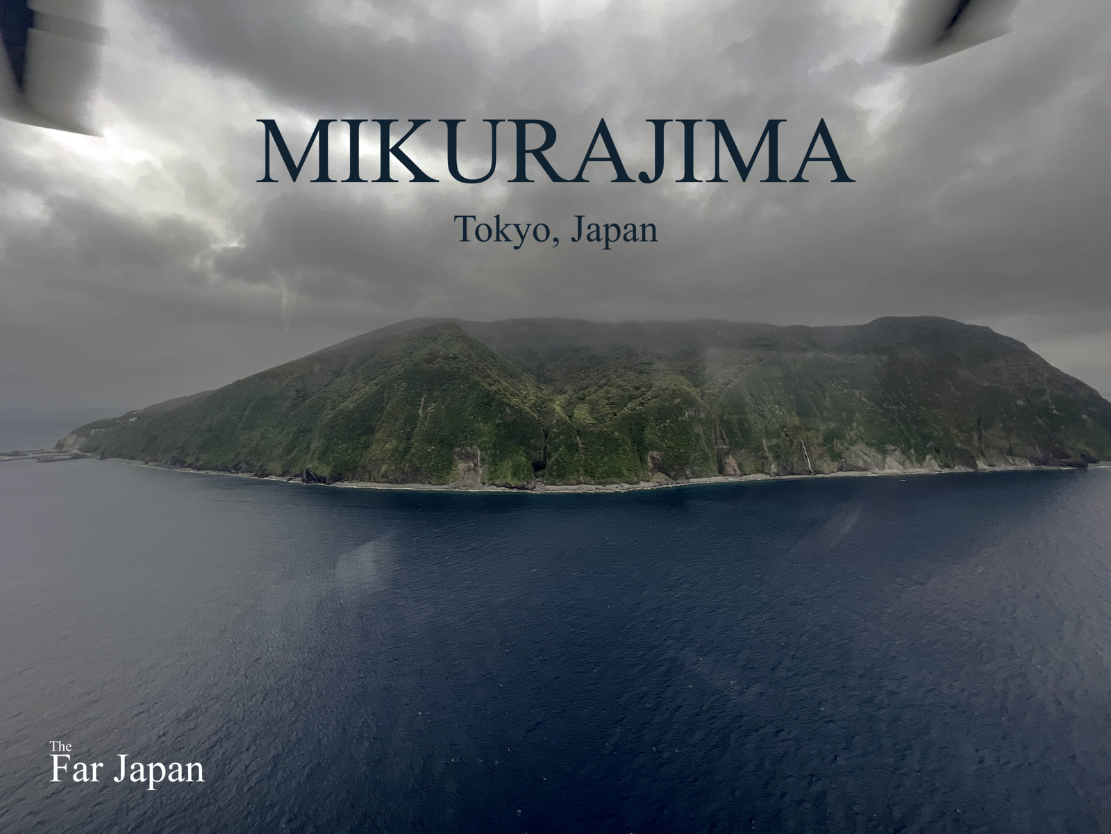 A dramatic aerial photograph looking down at the rugged, steep, heavily forested volcanic island of Mikurajima, surrounded by deep blue sea and under a dramatic, dark cloudy sky. The text "MIKURA JIMA" and "Tokyo, Japan" are overlaid at the top.